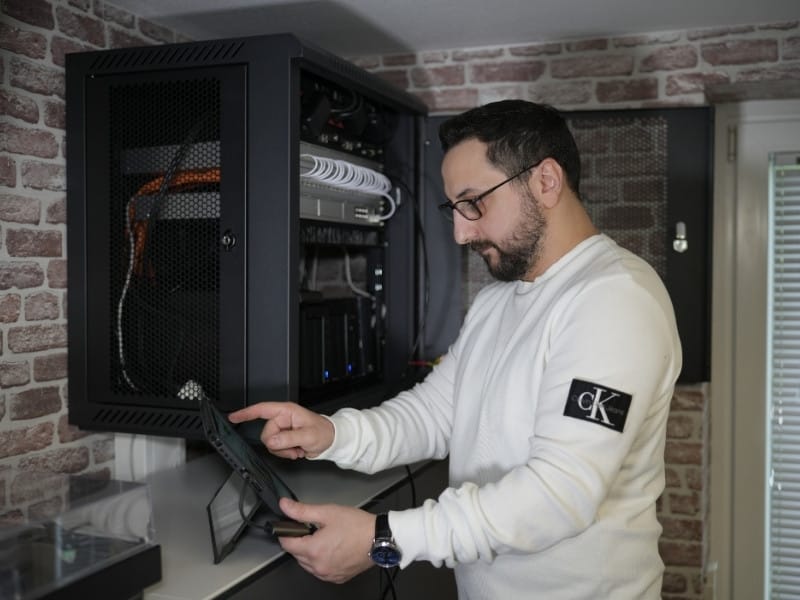 Kyrillos Poutakidis steht vor einem offenen, an einer Ziegelwand montierten Netzwerkschrank mit Switch, geordneter Verkabelung und einem Synology NAS-System, während er Einstellungen auf einem Tablet prüft. Das Bild zeigt die professionelle Einrichtung zentraler Datenspeicher durch syskypo für sichere IT-Dienstleistungen.